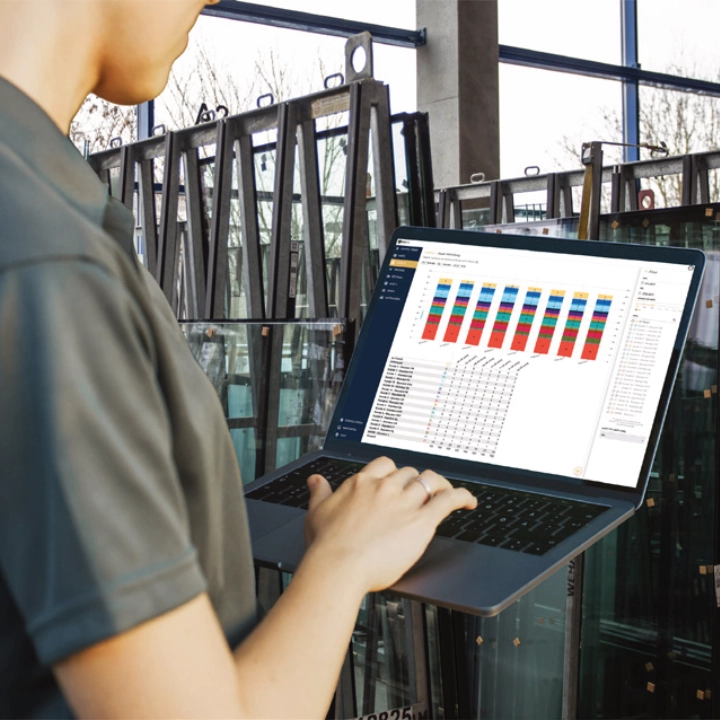 box-id_success-story_adamer-glas_production-hall Person arbeitet an einem Laptop mit Datenanalyse-Dashboard in einer industriellen Umgebung mit Glasscheiben im Hintergrund